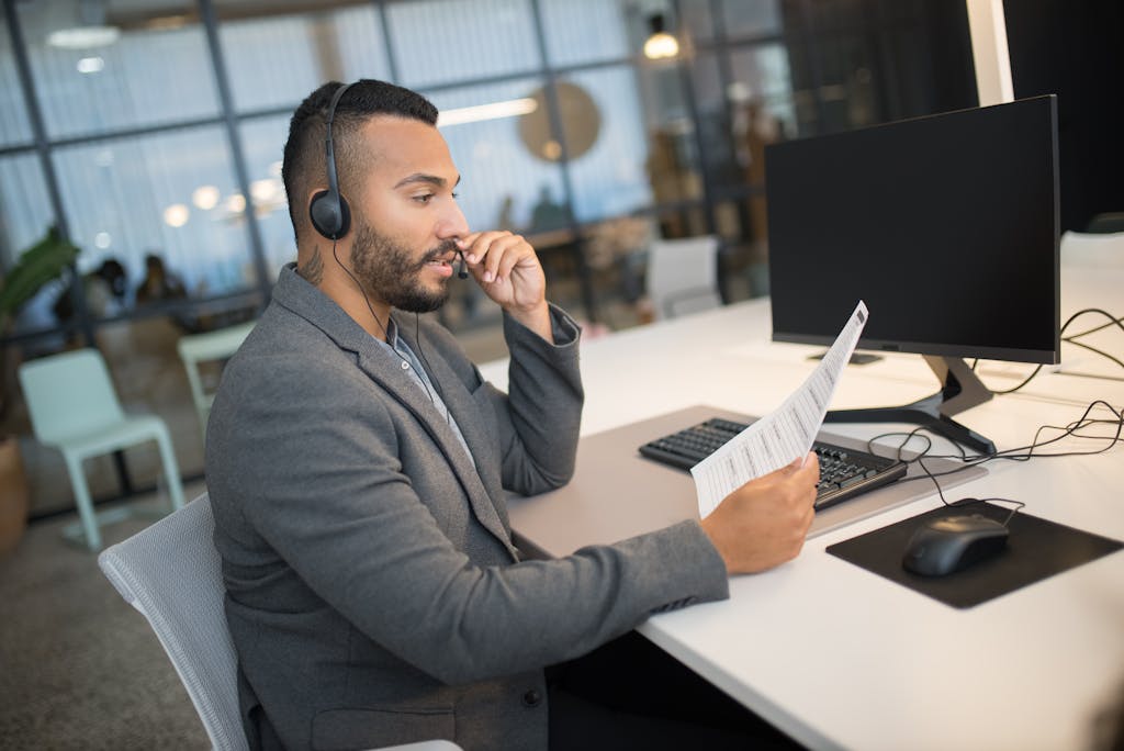 Young man in a call center, using a headset and reading documents at his office desk.